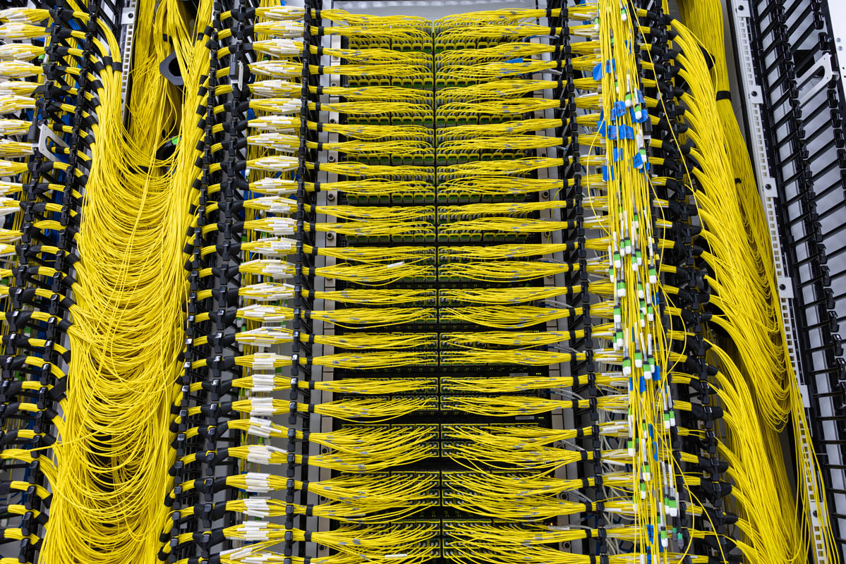 Rows of network switches at xAI’s Colossus data center, connected by dense, carefully routed yellow fiber-optic cables. Archive photograph from ServeTheHome’s 2024 tour.